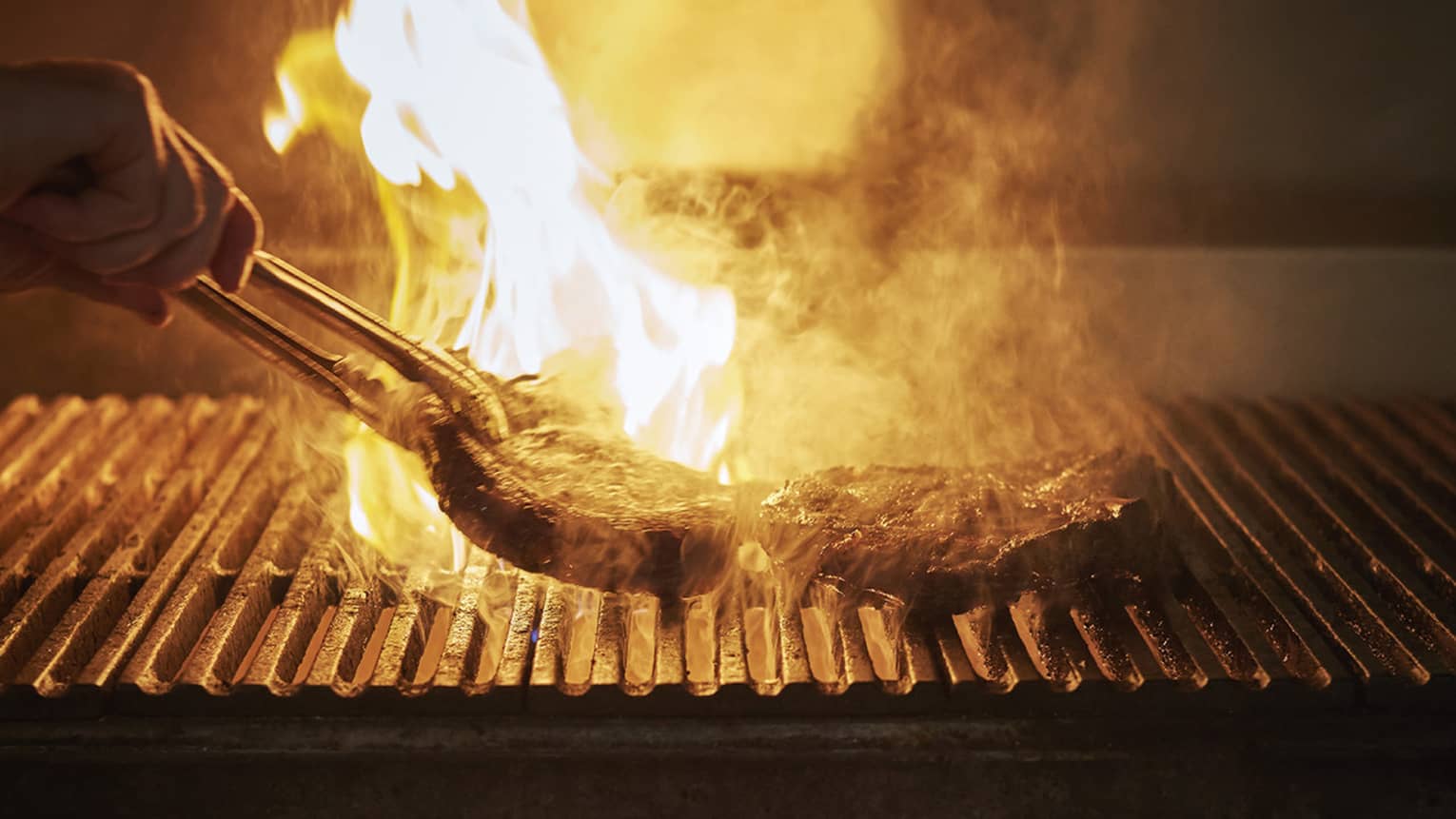 Fiery barbecue with silhouette of hand holding tongs, flipping large steak on grill