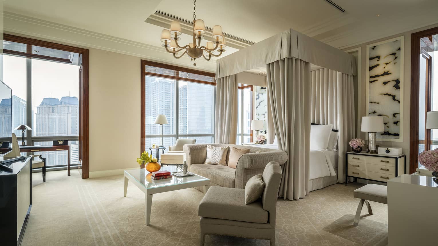 Hotel room canopy bed with white curtains behind sofa and seating area, large windows