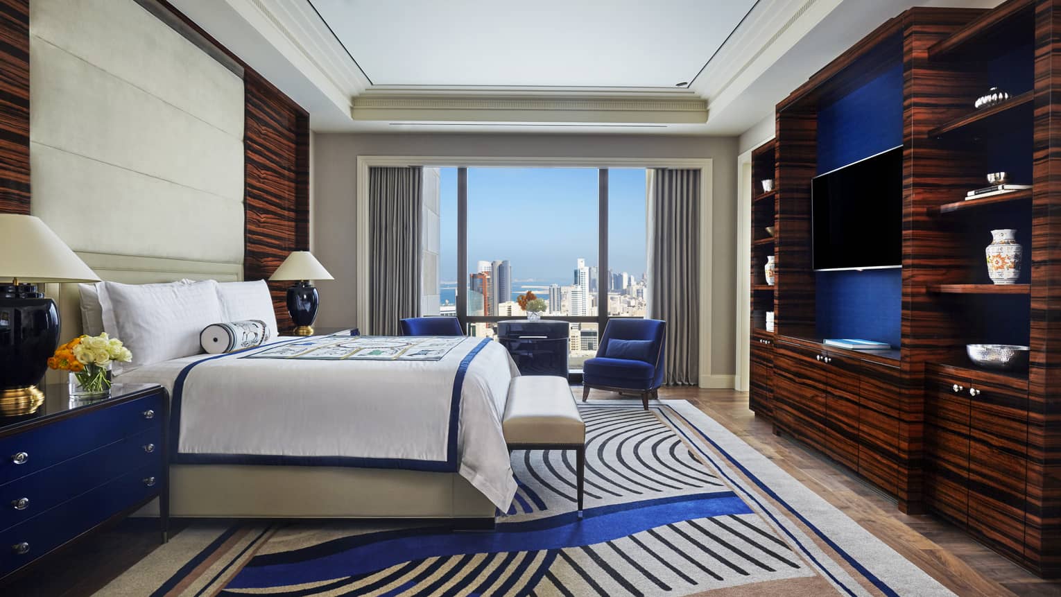 Hotel room with bed across from dark wood dresser and TV, blue accent chairs by window