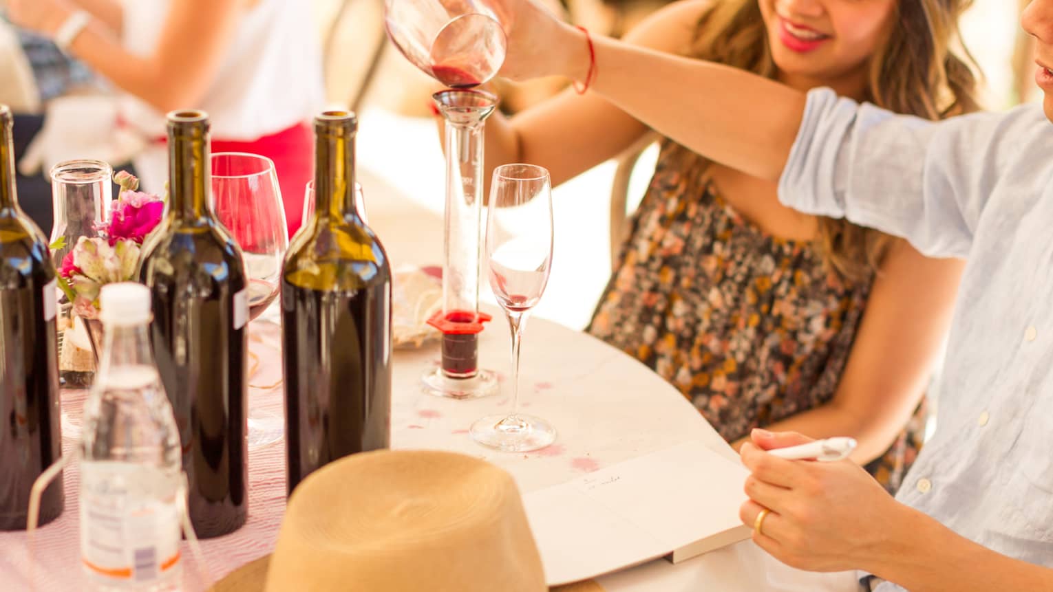 Man and woman pour red wine into glass tube on vineyard table with wine bottles, flowers