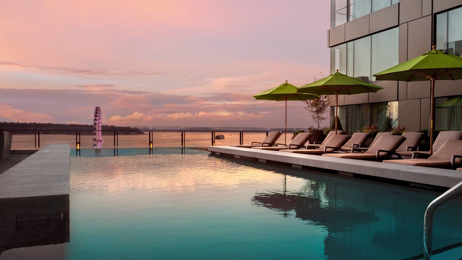 Pink sunset reflected on outdoor swimming pool lined with lounge chairs in front of bay, Seattle Wheel