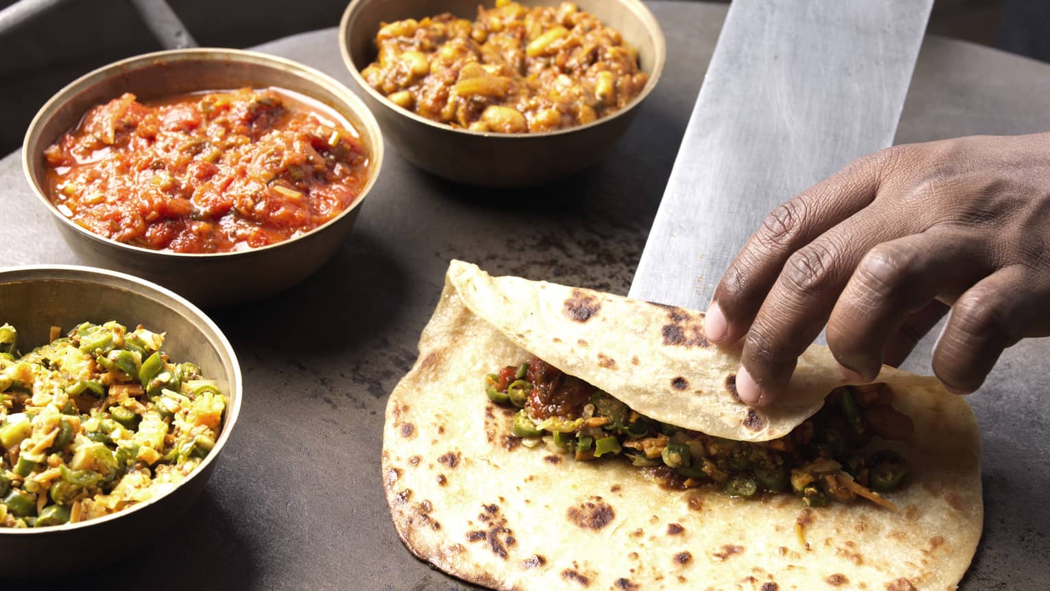 Hand sliding Mauritian Farata flatbread with filling onto counter, three bowls with curries