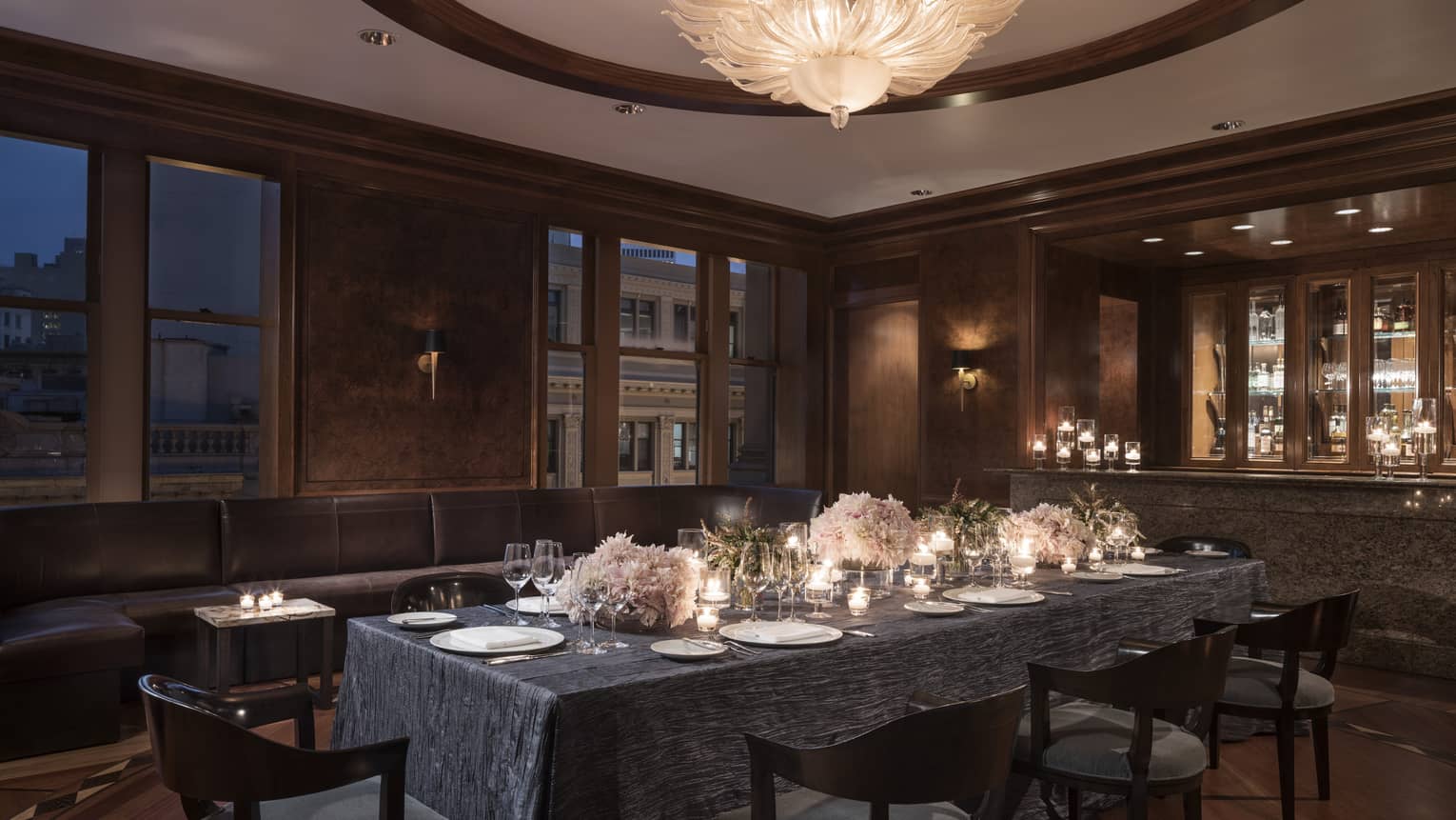 Private dining table with candles, flowers near long leather banquette, windows at night