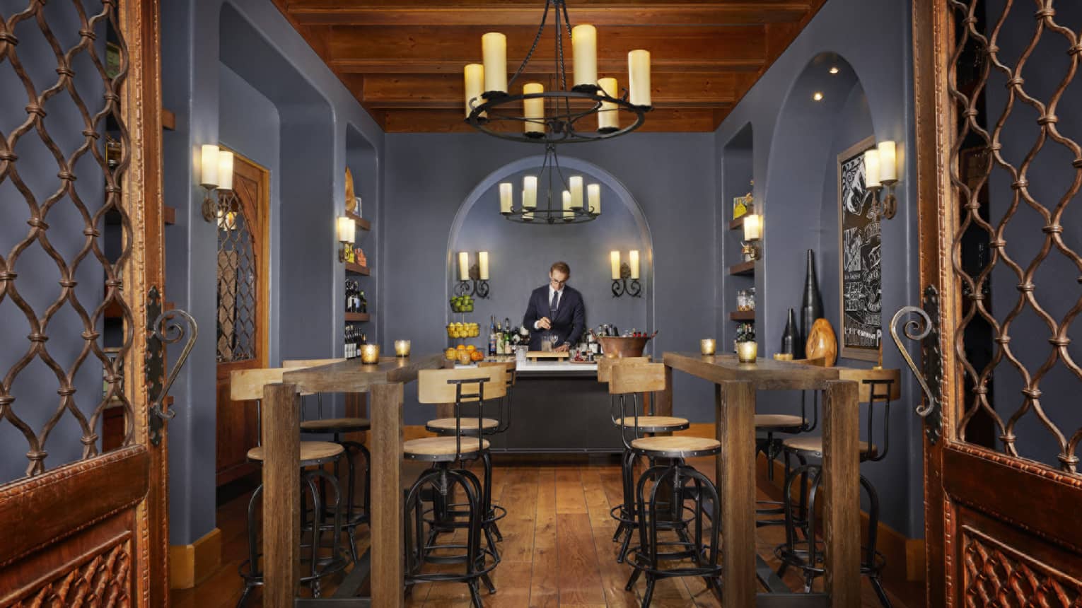 Talavera bartender under arched wall, chandeliers with tall candles behind wood bar tables