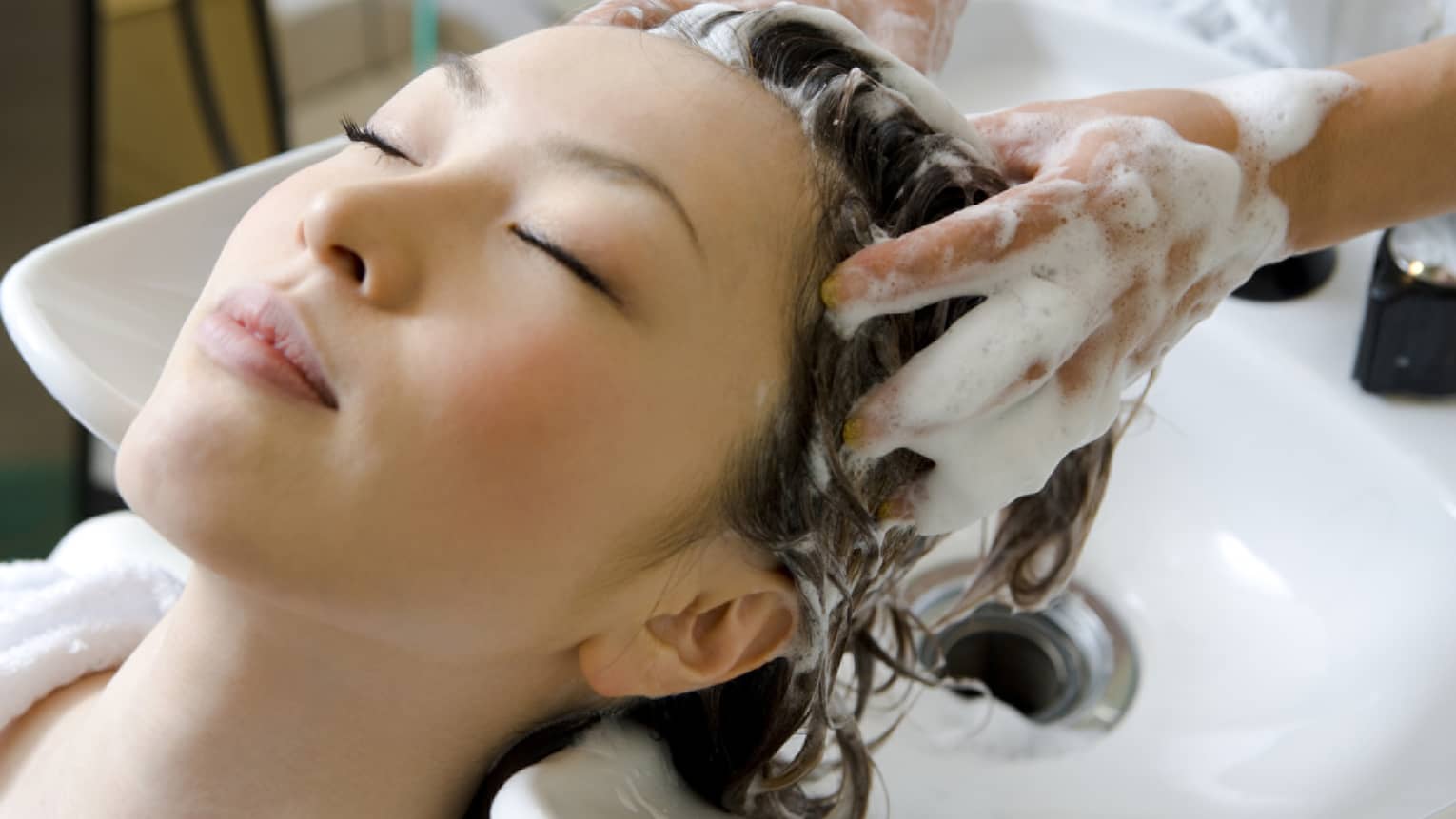Woman closes eye and leans back against sink as hands massage shampoo into her scalp