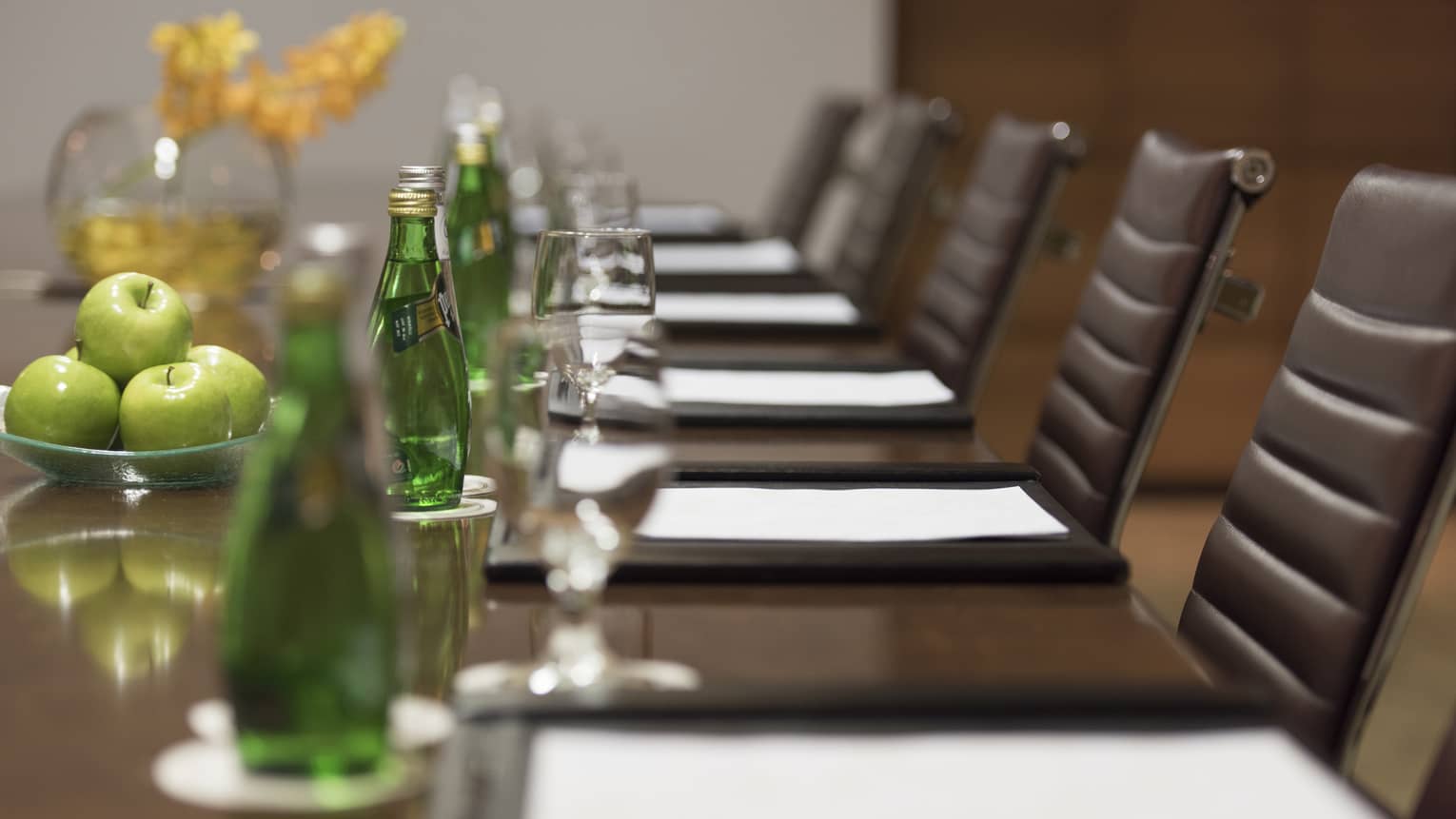 Close-up of agendas in front of leather chairs at long meeting table with green glass bottles, apples 