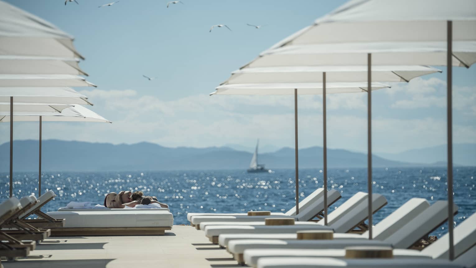 A sail boat sails in the distance while white lounge chairs are ready for guests and lined up next to the oceanside Nafsika Pool in Athens. 