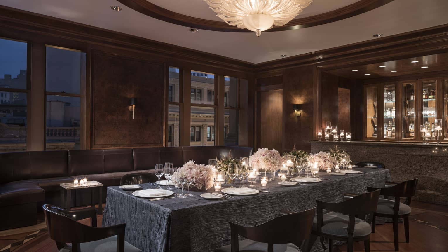 Private dining table with candles, flowers near long leather banquette, windows at night