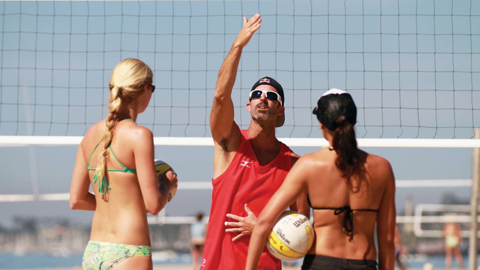 Two women in bikinis hold volleyballs as man demonstrates serve in front of net