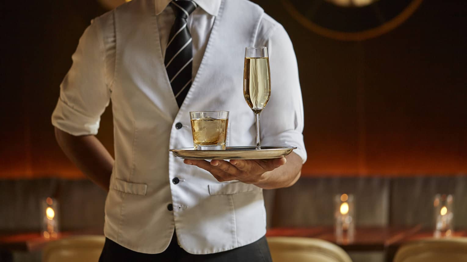 Lounge server wearing white vest holds tray with liquor in rock glass, Champagne in flute