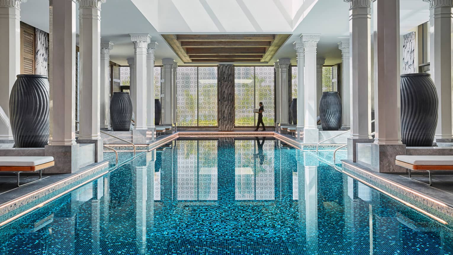 Hotel staff walks by window at end of long indoor swimming pool lined with pillars, large vases