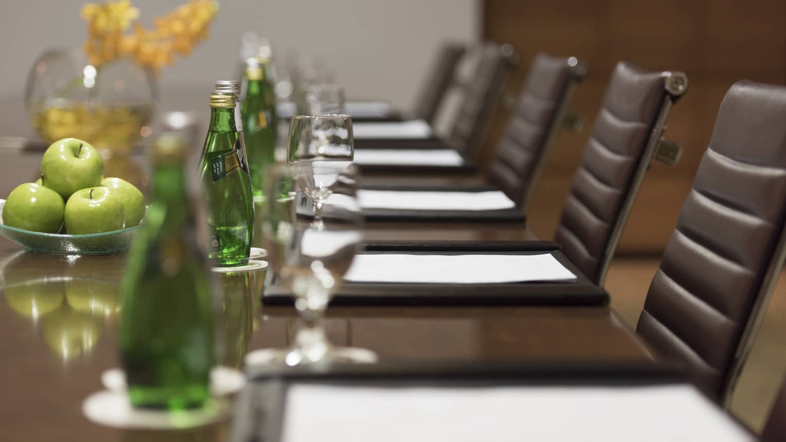 Close-up of agendas in front of leather chairs at long meeting table with green glass bottles, apples 