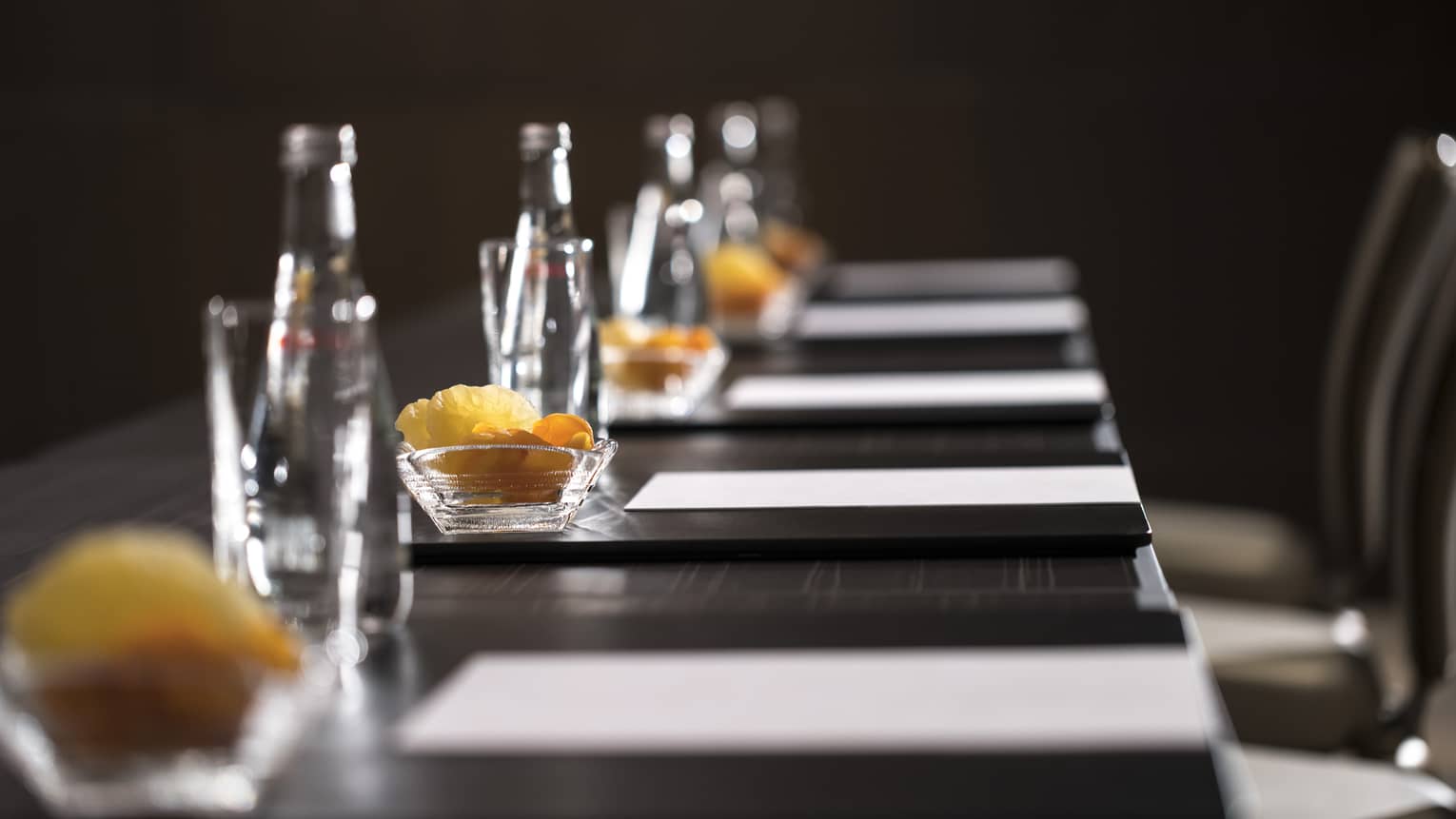 Close-up of boardroom meeting table with row of agendas, glass water bottles, fruit slices in bowls