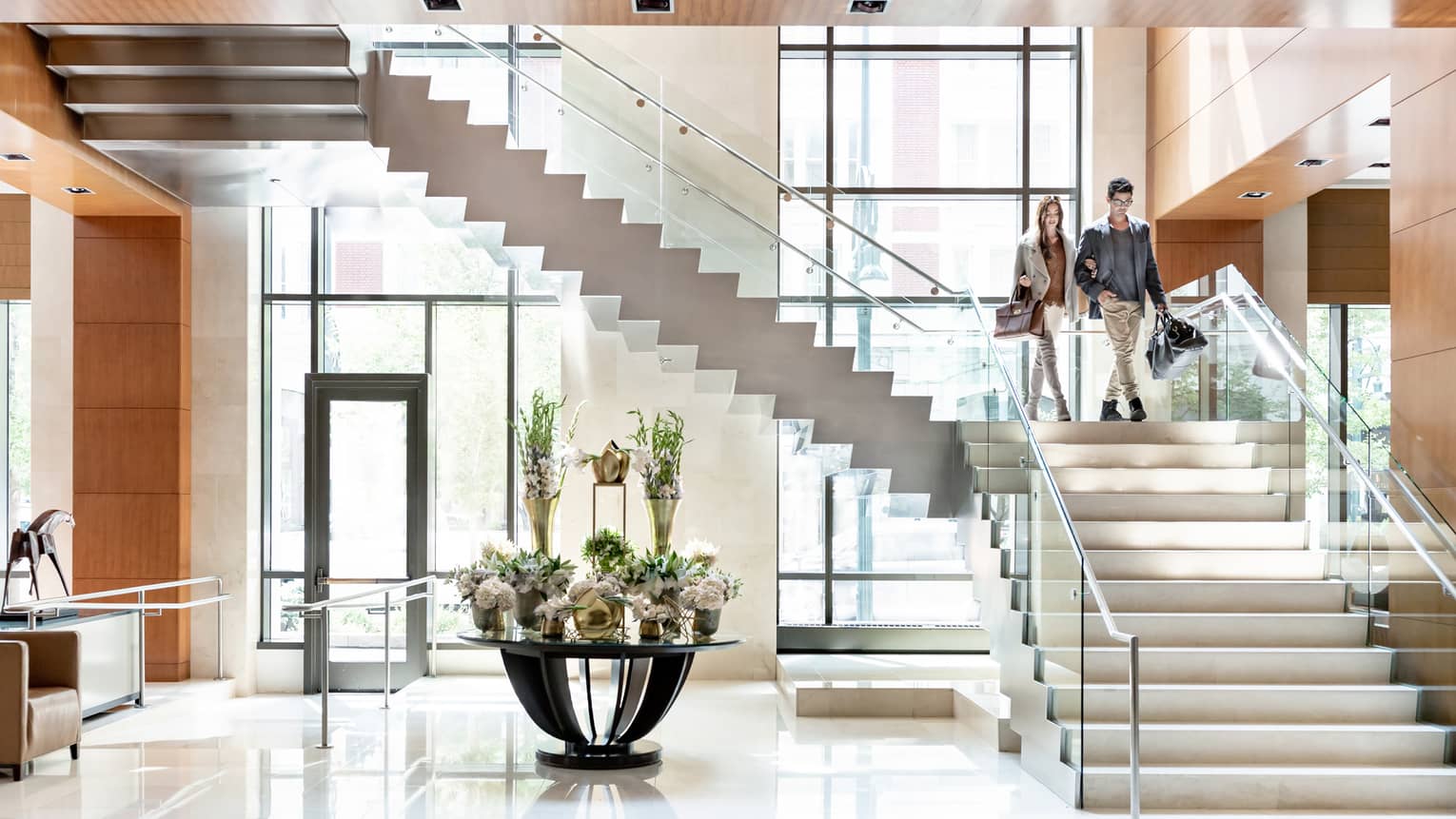 Couple walks down modern staircase with glass railings to hotel lobby with flowers on table