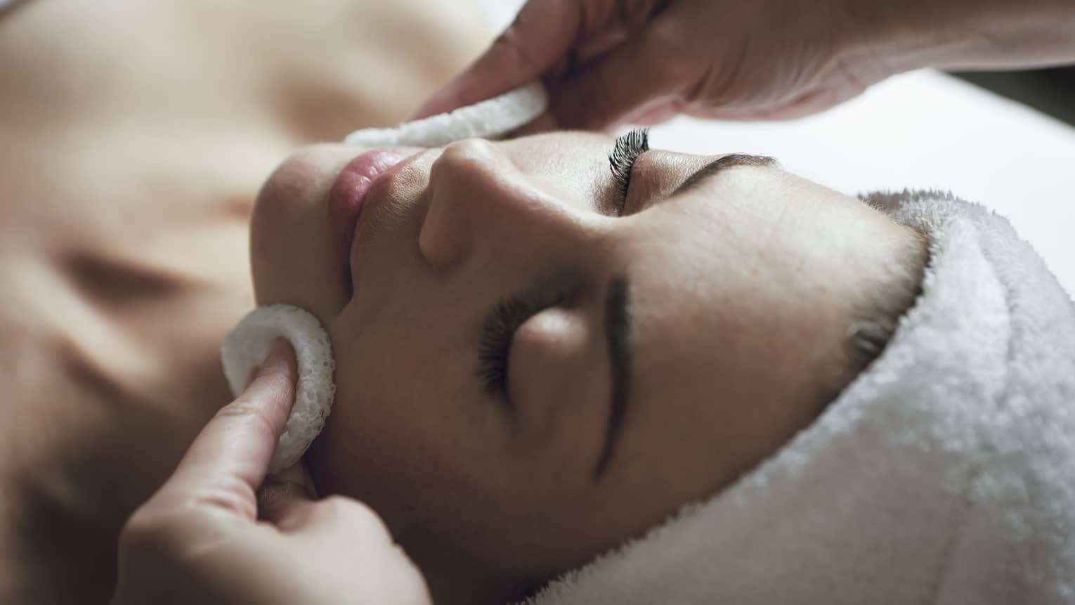Spa staff applies facial cleanser to woman's face, her eyes closed and towel around hair