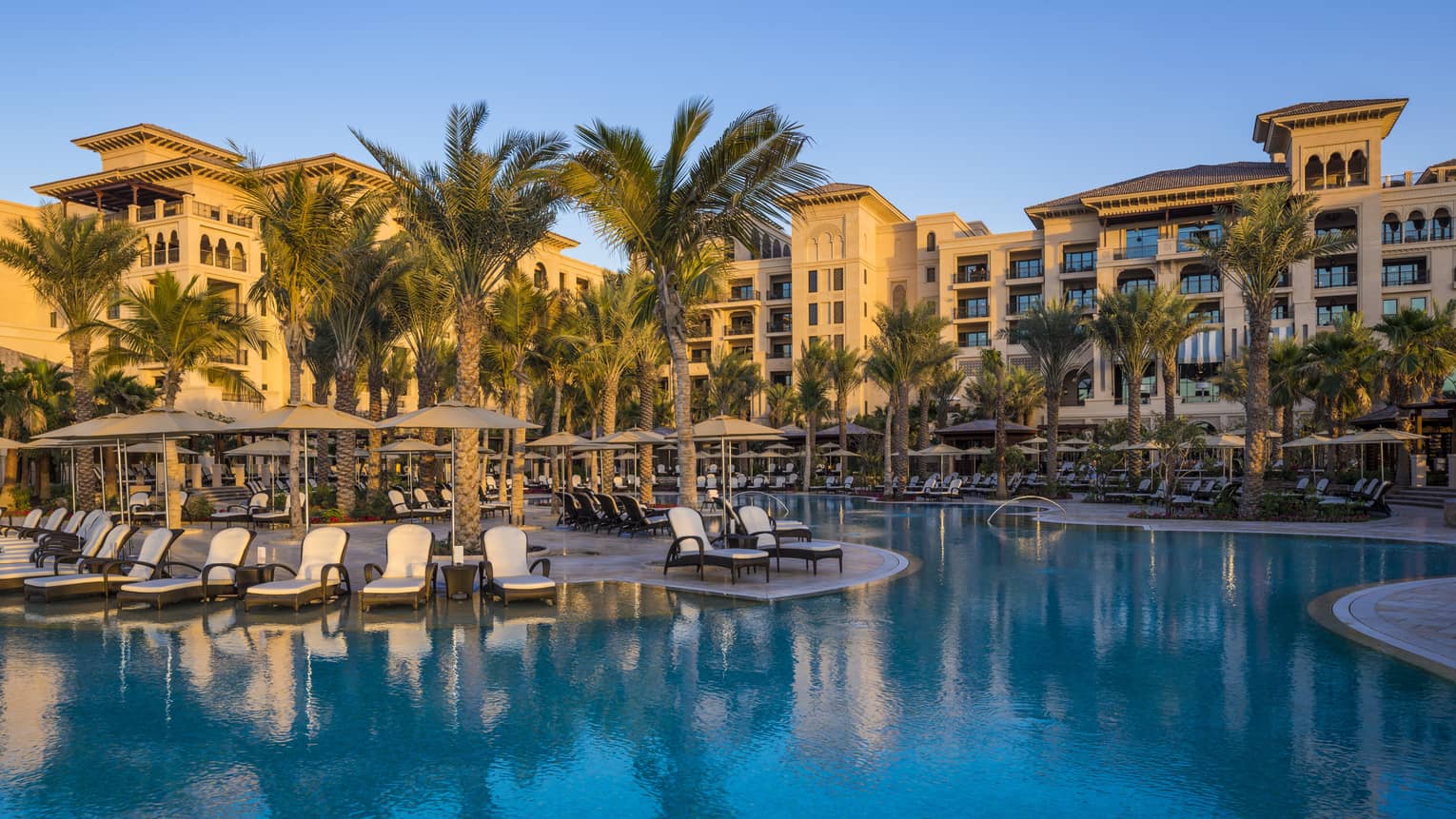 Winding blue outdoor swimming pool lined with white chaise lounge patio chairs, palm trees