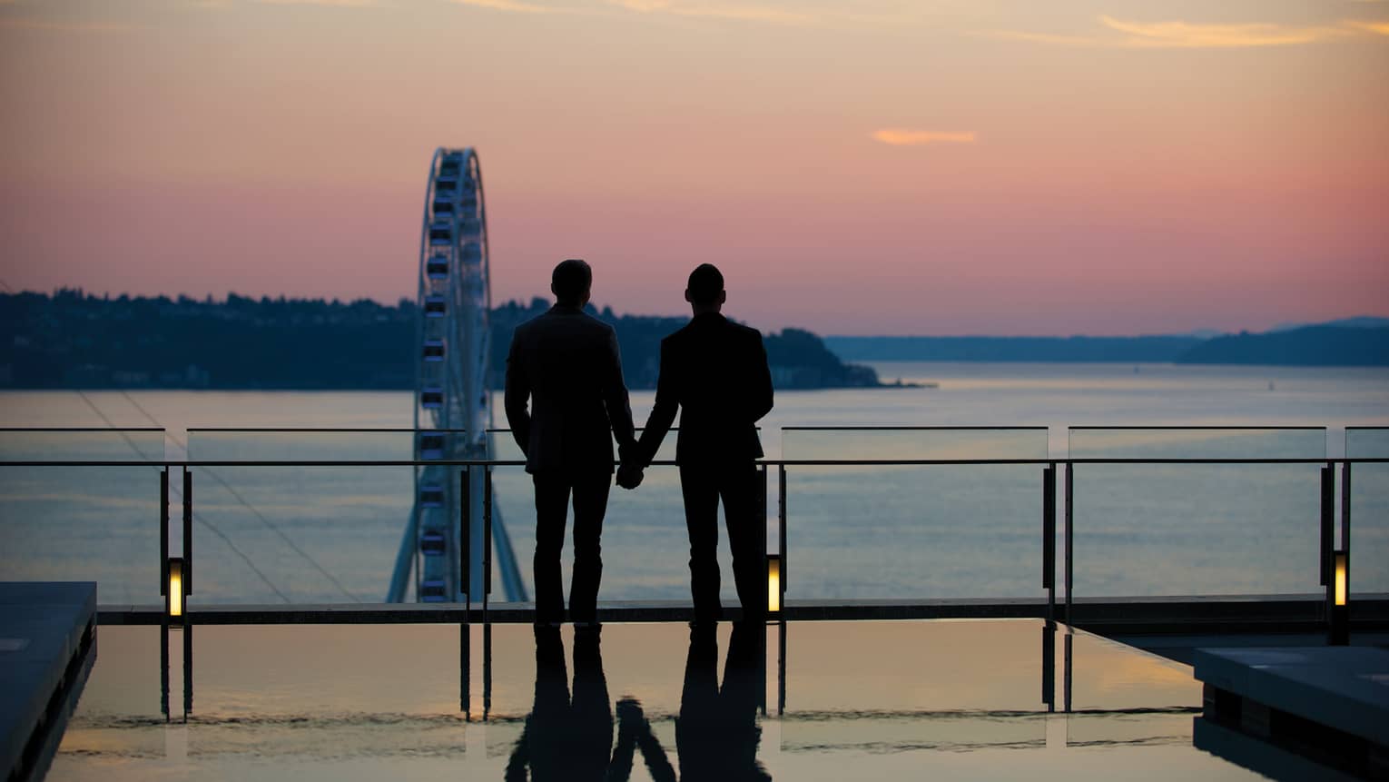 Silhouette of couple wearing suits and holding hands, looking over ocean at sunset