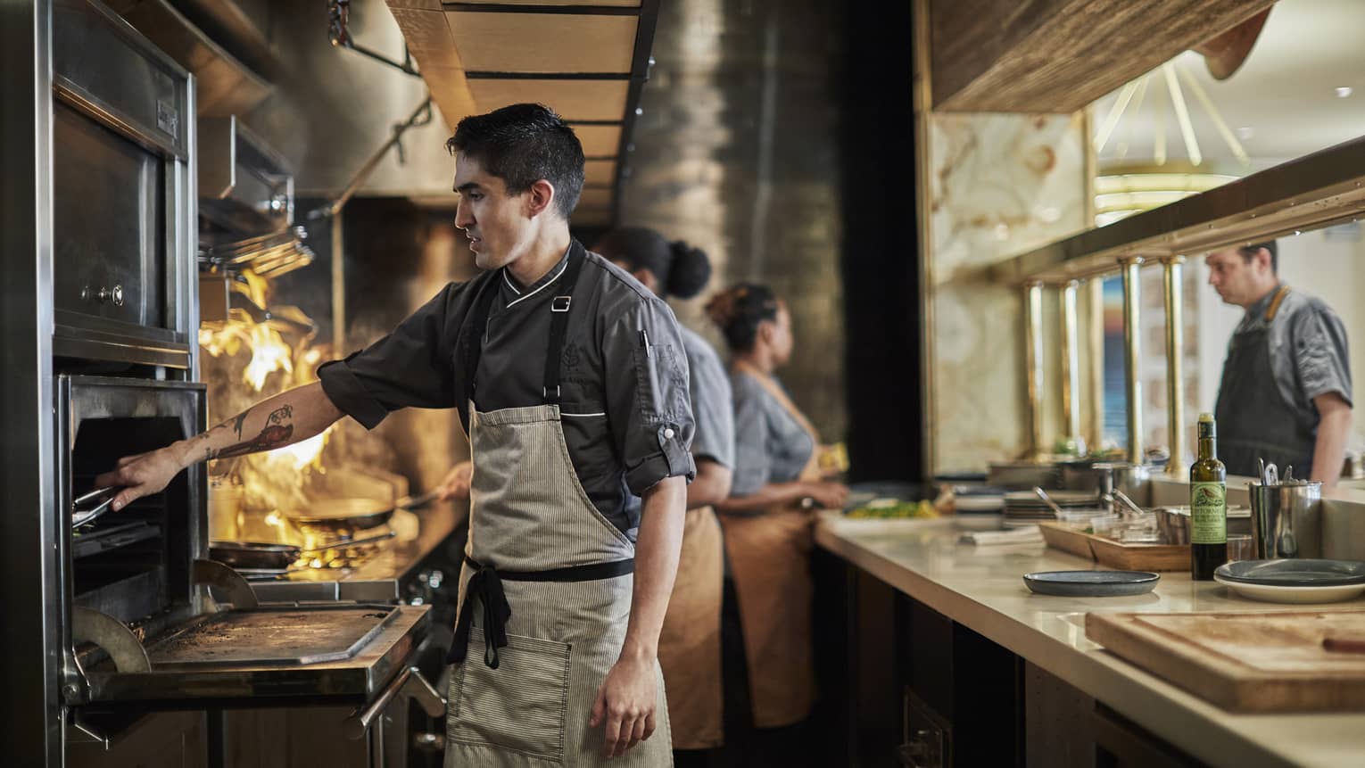 Esquilina Chef with tongs reaches into oven near flaming stove, kitchen staff in background