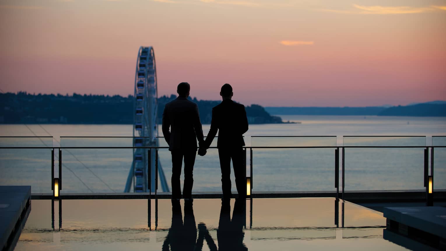 Silhouette of couple wearing suits and holding hands, looking over ocean at sunset