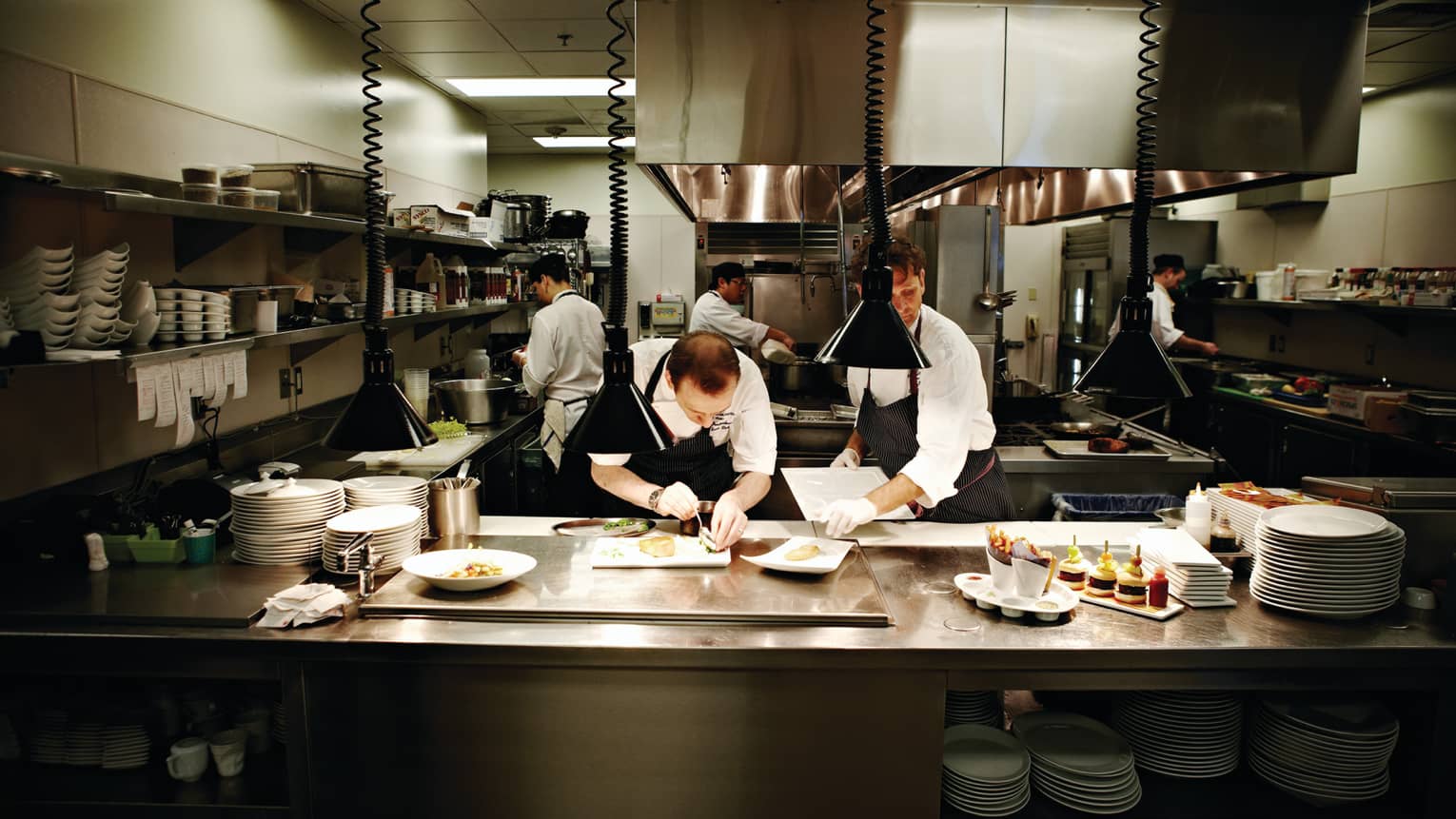 Two chefs plate gourmet dishes on kitchen line as cooks work in background