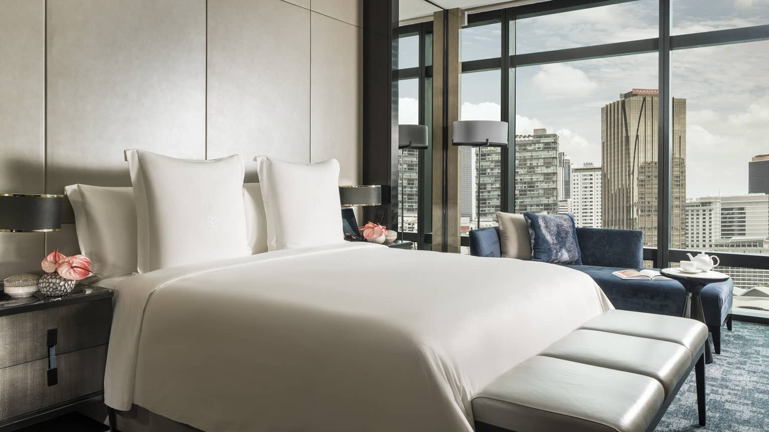 Hotel bed with white linens, modern bench at foot, chaise by floor to ceiling window