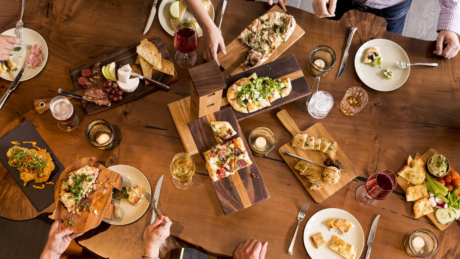 Aerial view of people dining around rustic wood table with platters of flatbreads, appetizers