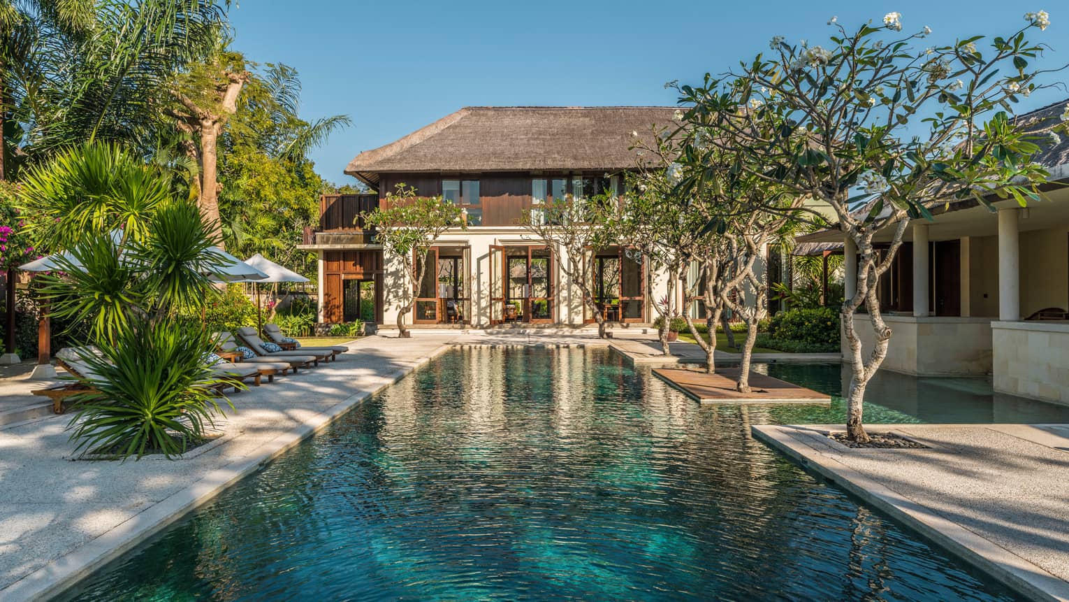 Two-storey villa at end of long outdoor swimming pool lined with trees, tropical plants