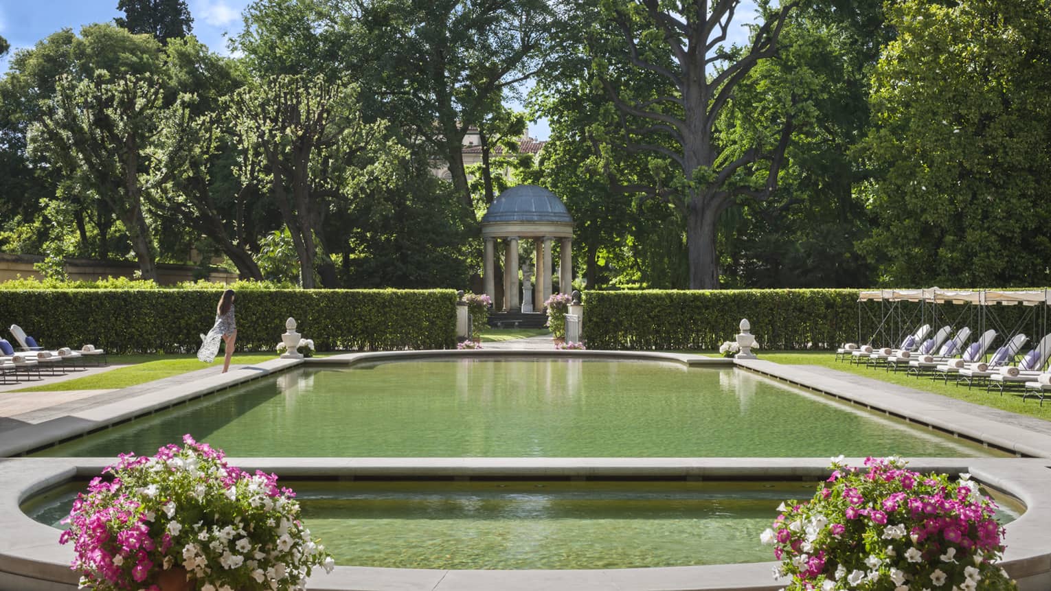 Woman in flowing robe walks past stone ledge of outdoor swimming pool surrounded by gardens