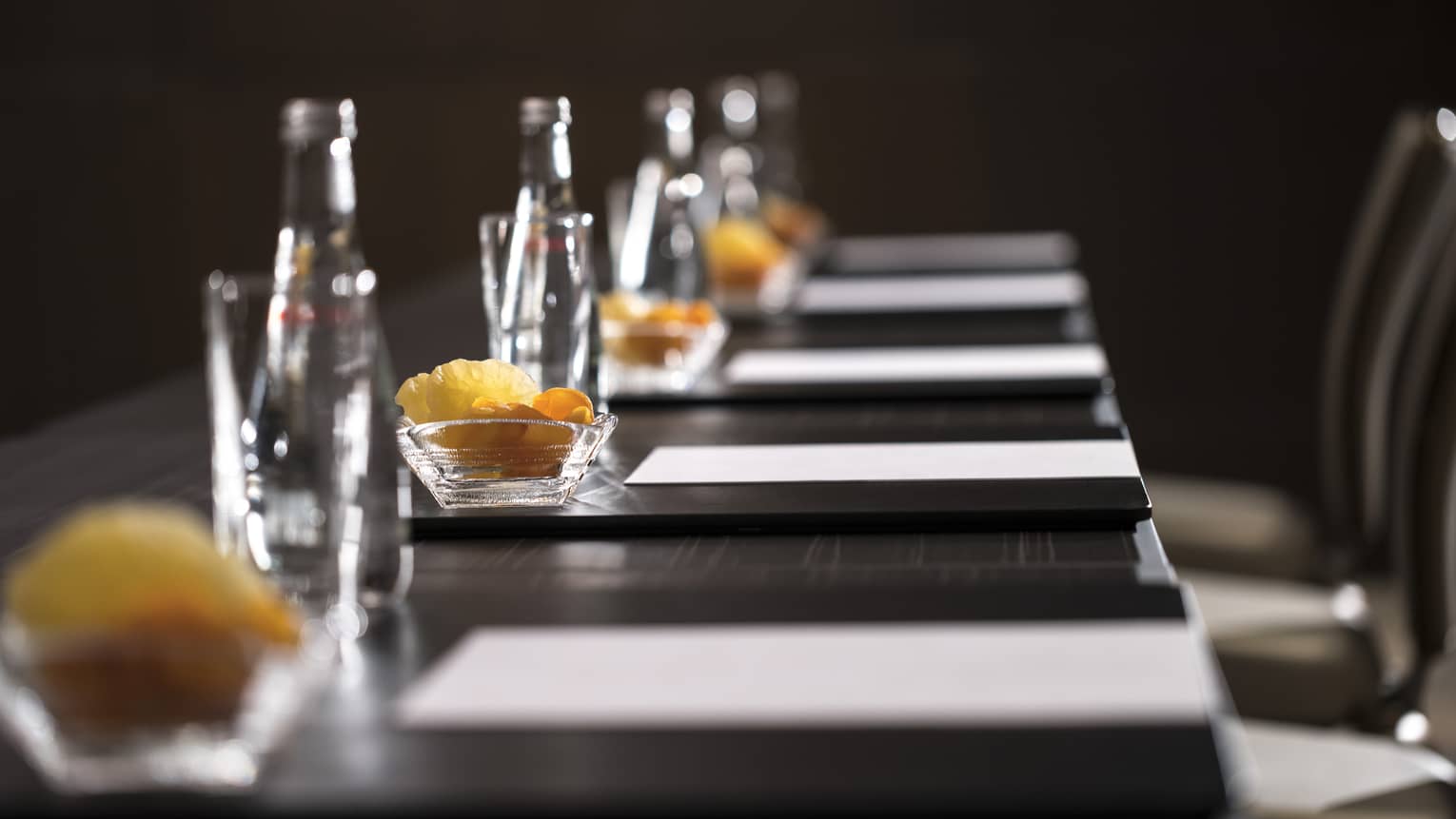 Close-up of boardroom meeting table with row of agendas, glass water bottles, fruit slices in bowls