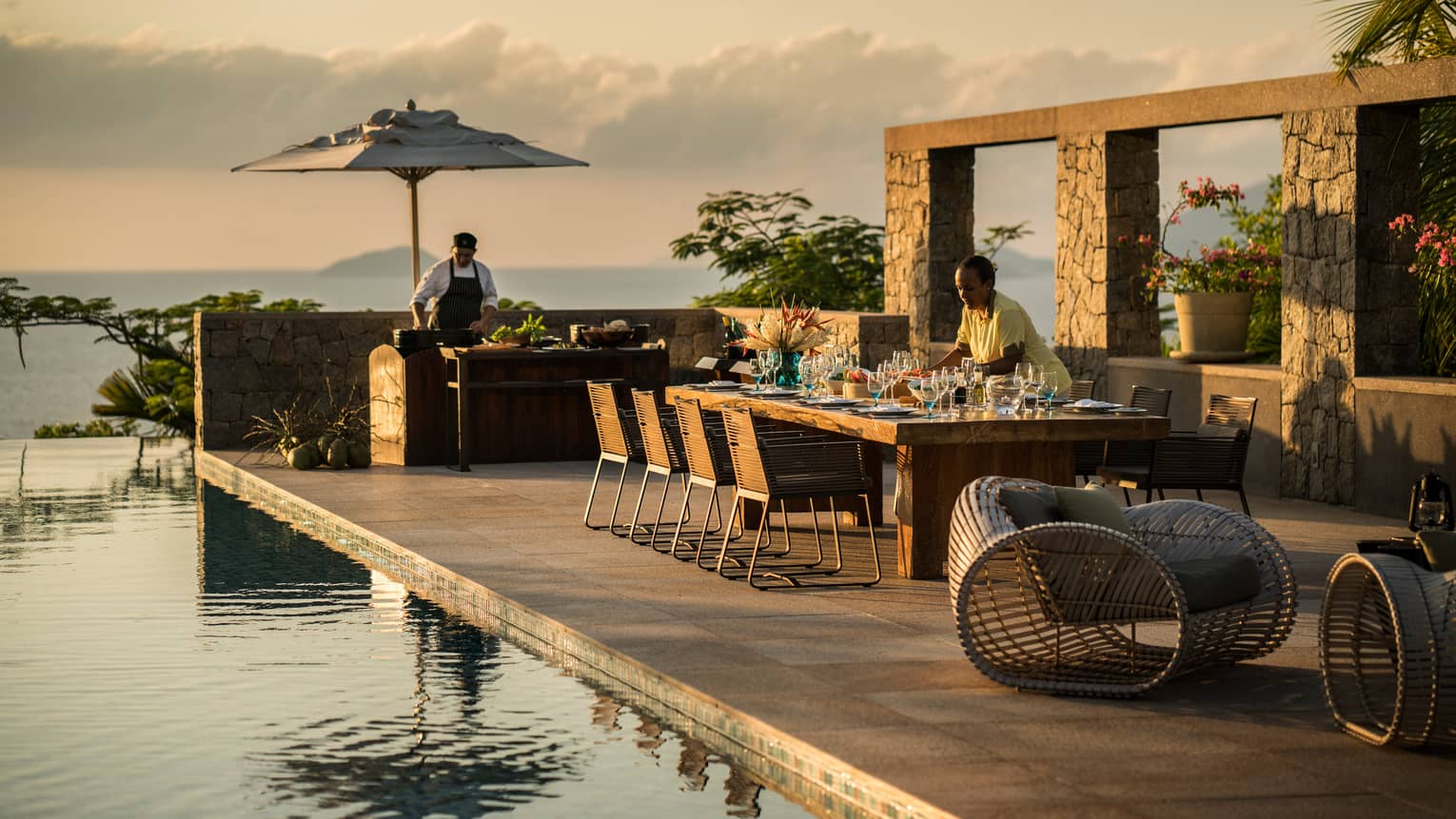 Residence Villa close-up of server setting long dining table by swimming pool at sunset 