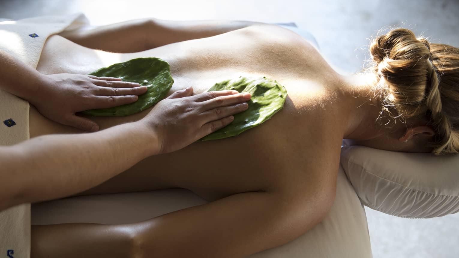 Woman face-down on table as masseuse rubs Nopal cactus leaves on her back with Mexican Nakieri Treatment