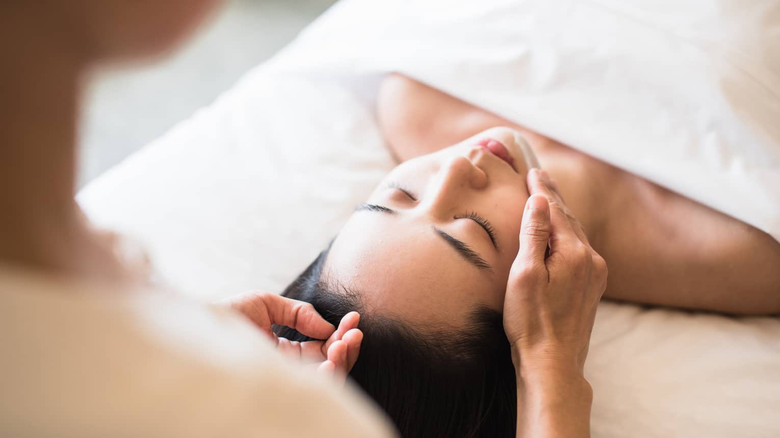 Woman lying under white sheet on massage table, eyes closed as spa staff rubs lotion on her chin