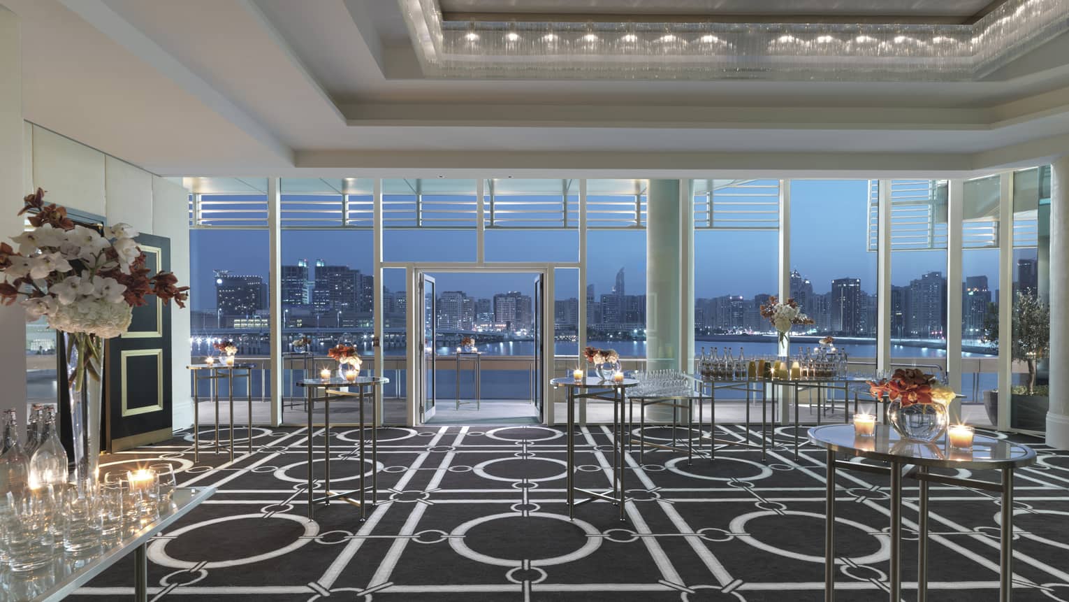 Cocktail reception tables with small candles, flowers in Liwa Ballroom, floor-to-ceiling windows with sky at dusk