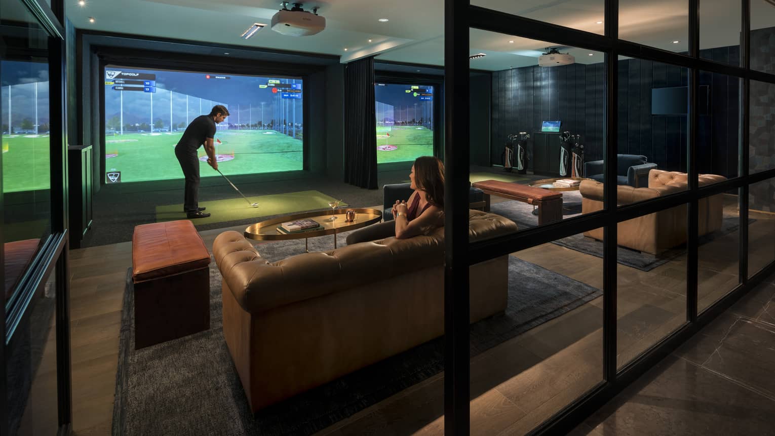 Topgolf lounge, man swings club by large golf simulator screen, woman sits on leather sofa behind him
