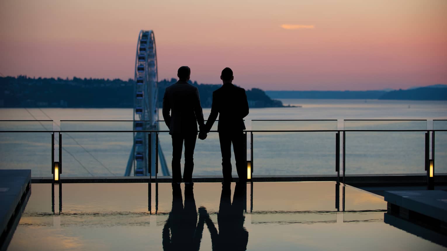 Silhouette of couple wearing suits and holding hands, looking over ocean at sunset