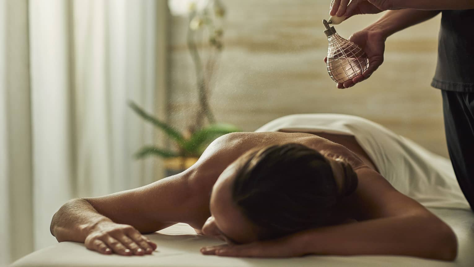 Spa staff sprays perfume bottle over woman lying on massage table