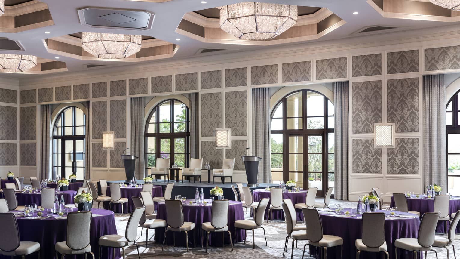 Grand Ballroom with round banquet tables under geometric chandeliers, sunny arched windows 