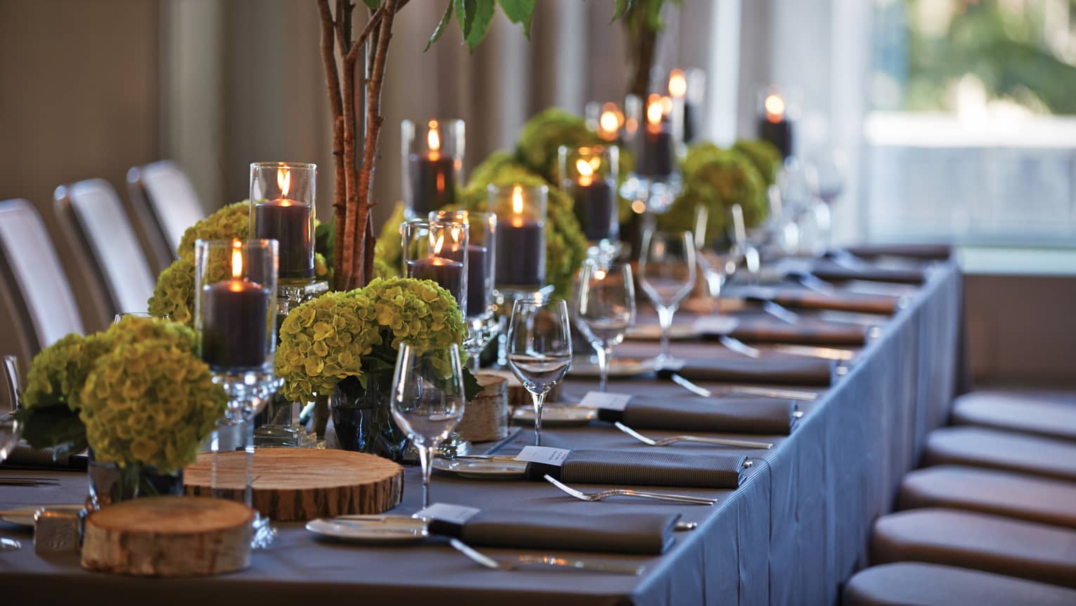Close-up of long banquet table set with wine glasses, rustic wood platters, green flowers, candles
