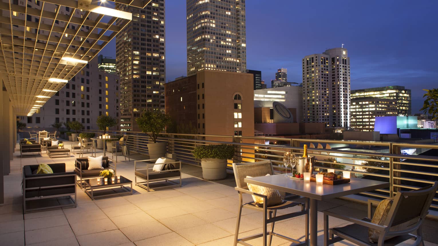 Large rooftop patio at night with candle-lit tables, champagne on ice, black cushioned chairs, city lights