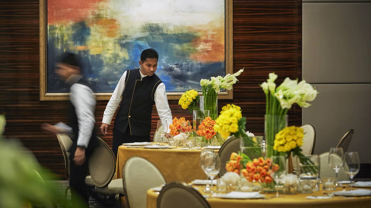Hotel staff sets round formal dining table with yellow linen, vases of fresh orange, yellow flowers