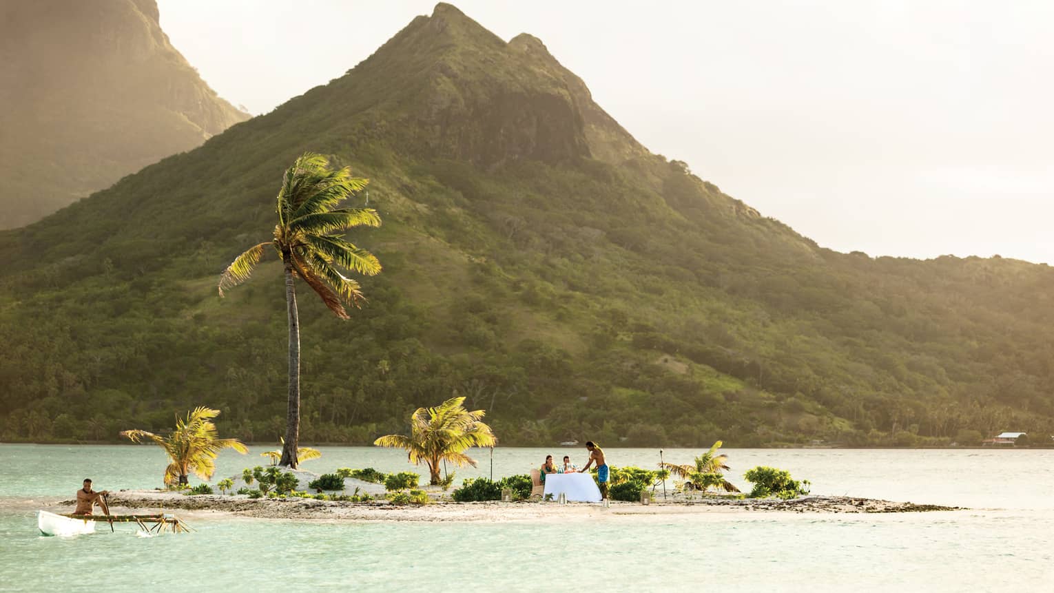 Couple dines on small private island with windswept palm trees in lagoon under mountain