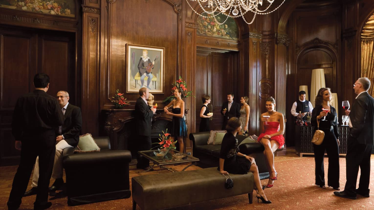 People wearing cocktail attire socialize in elegant ballroom with wood walls, crystal chandelier, leather benches 