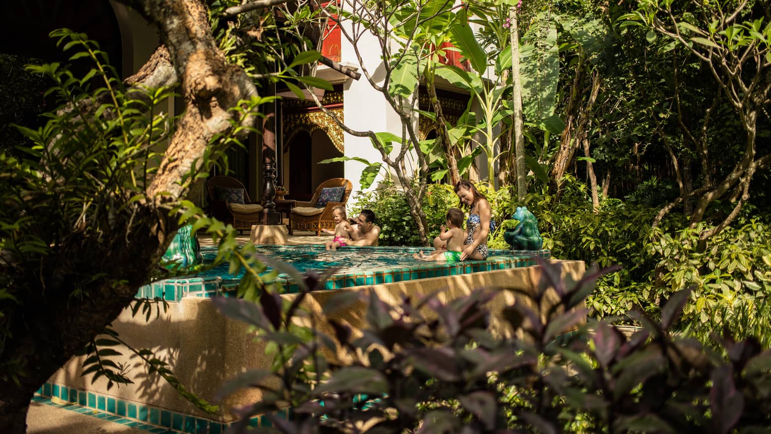 Couple with two babies sit on edge of private pool in front of bungalow by garden