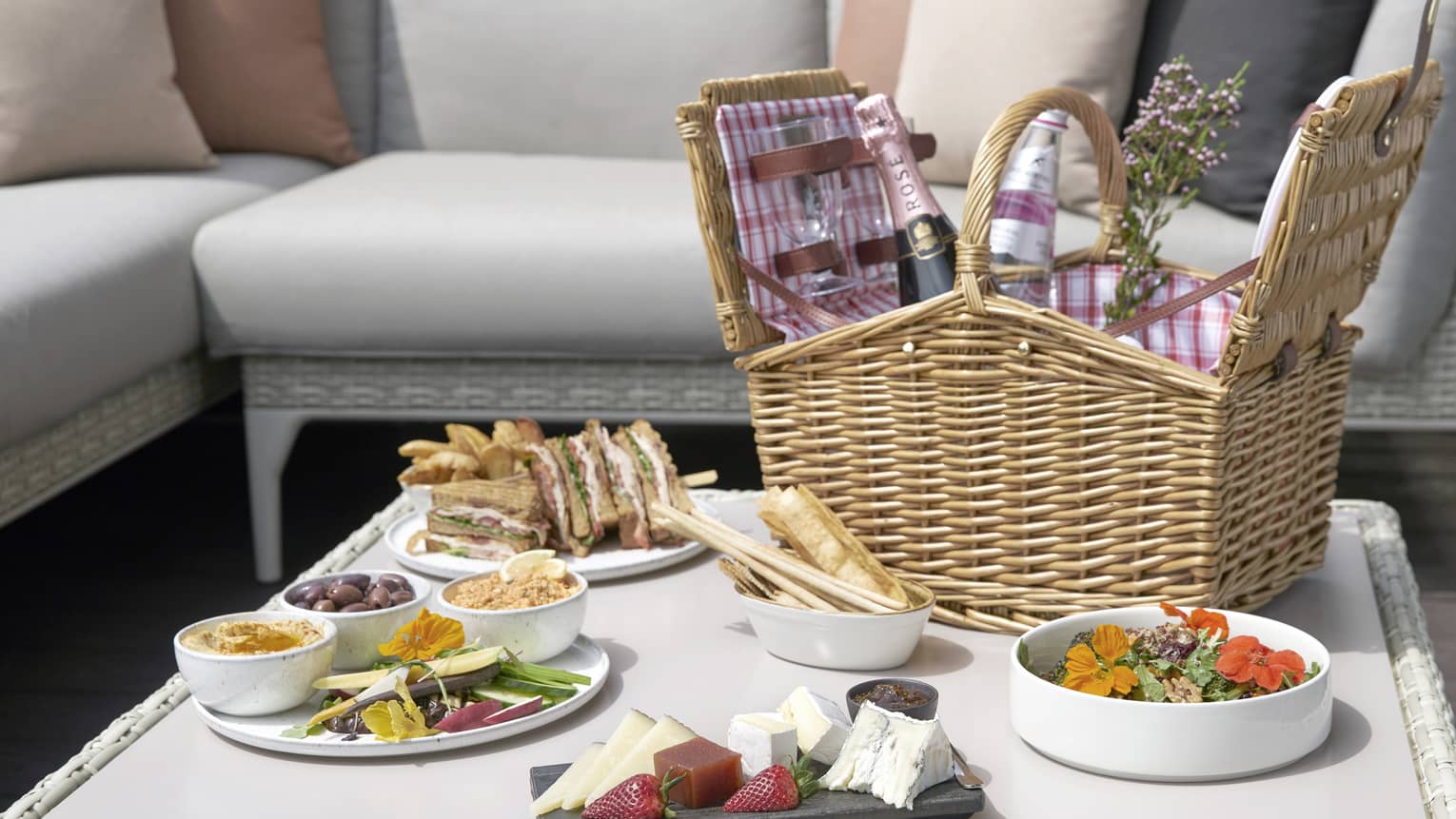Patio lounge table with wicker picnic basket with Champagne bottle, plate with clubhouse sandwich, appetizer platters