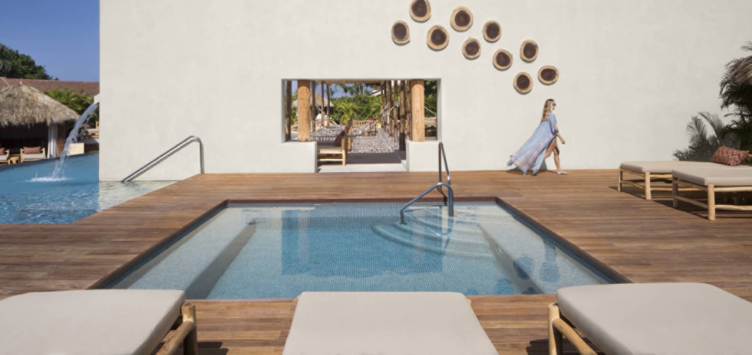 Woman in flowing blue beach robe walks past square plunge pool on modern deck near lounge chairs, hammock