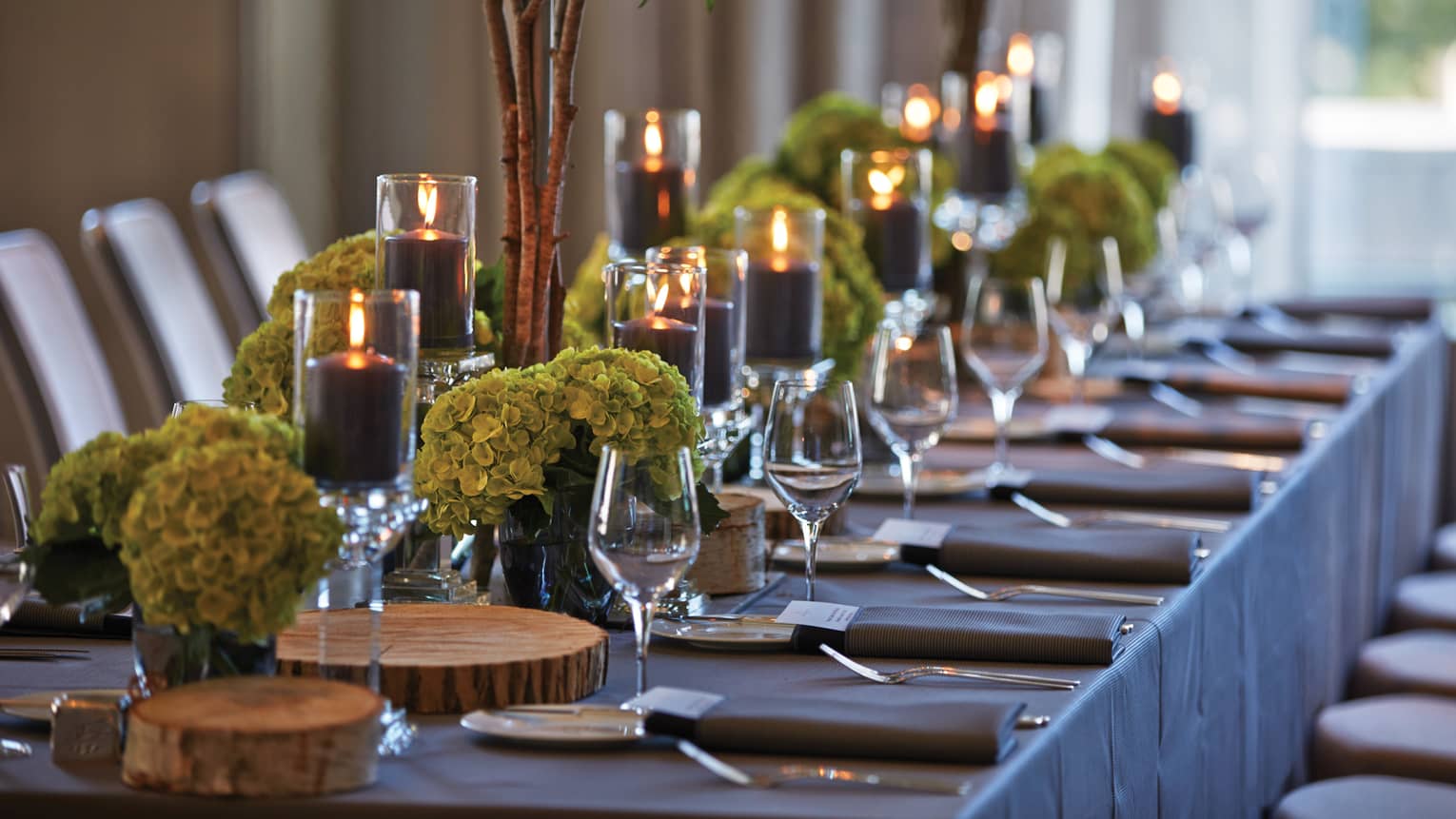 Close-up of long banquet table set with wine glasses, rustic wood platters, green flowers, candles
