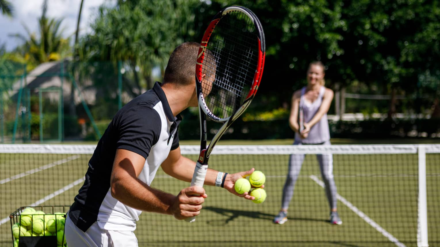Couple playing tennis on court, man prepares to serve tennis balls to woman ready to swing