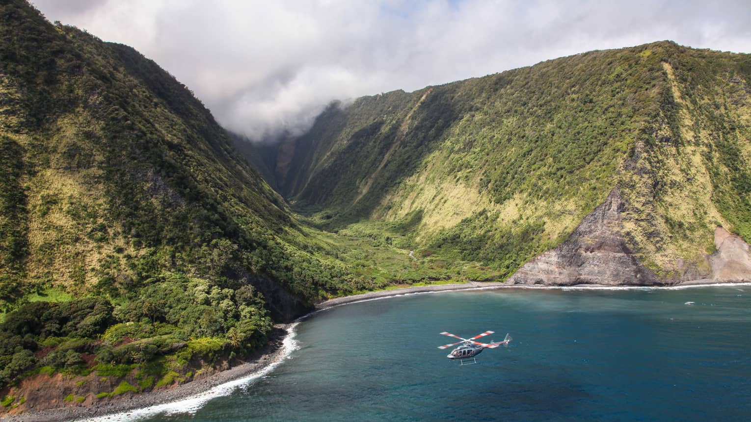 Helicopter flying over ocean near sweeping green volcanic mountains