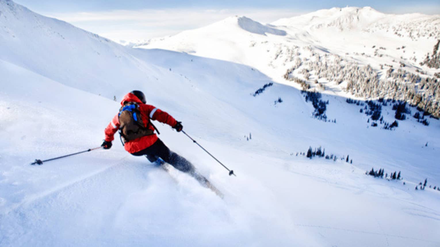 Back view of skier in red jacket, backpack, skiing down mountain slope on sunny day