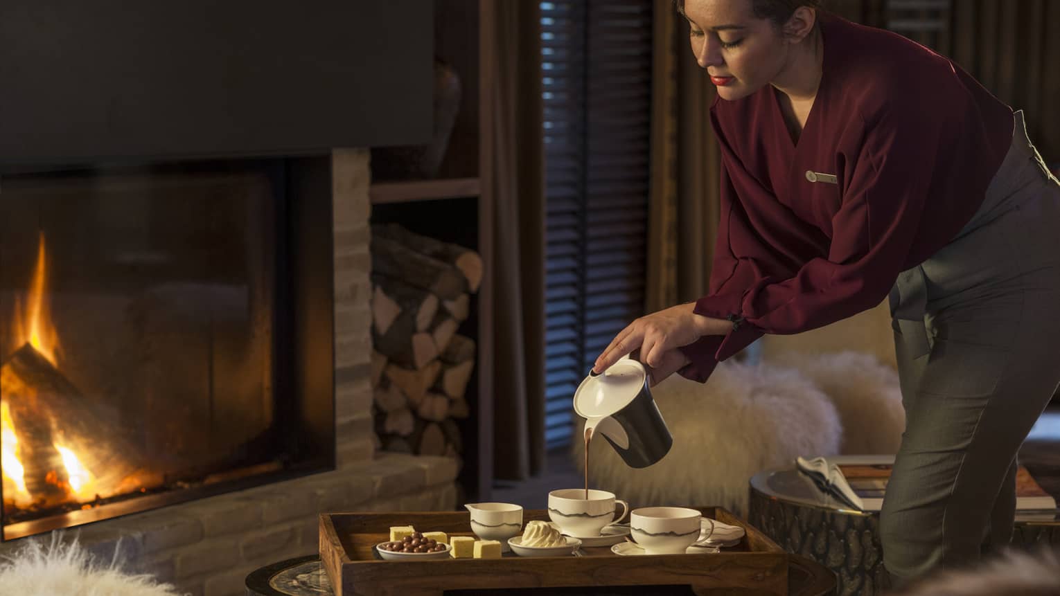 Hotel staff pours hot chocolate into modern cups on tray by large indoor fireplace, flames 